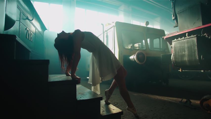 Woman in white dress posing on stairs surrounded with trains 4K. Low angle wide view of person in focus lying on stairs inside a hall of smoke and sun rays.