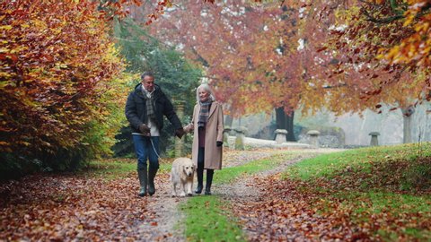 Happy Retired Senior Couple Taking Dog For Walk Along Path In Autumn Countryside Together