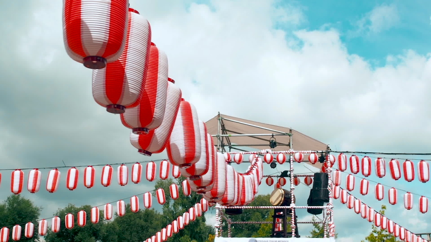 The stage of the Yagura with a big japanese taiko drum Odaiko. Many paper red-white lanterns Chochin swinging in the wind on white clouds background. Scenery for the holiday