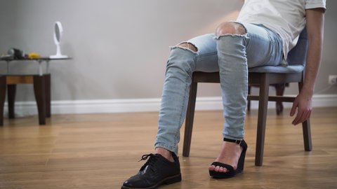 Unrecognizable Caucasian man in casual clothes sitting on chair with one foot in men's boot and other one in women's high heel shoe. Transgender, intersex people, self identification.
