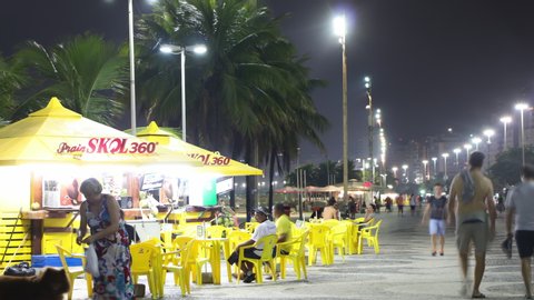Rio de Janeiro, Brazil - January 18, 2020: Night Timelapse of street bar next to Copacabana beach with people chatting and partying. View of plastic table and chair, road traffic behind.
