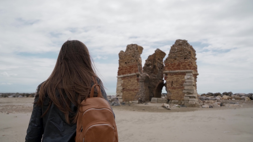 Long-hair brunette girl with a backpack approaches ruins of an ancient fortress Torre Flavia in Ladispoli, Italy. Rear view.
