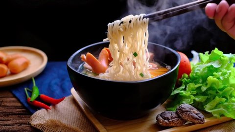 Man's hand pick up steamy instant noodle /hot ramen with chopsticks with ingredients on wood table. Hot ramen is delicious & cheap. Quick meal, instant noodle and cheap Asian steamy food concept. 