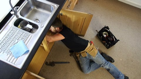 Plumber / Handyman fixing a leak. The tradesman is lying down under the sink to repair the pipe. Camera Crane shot.