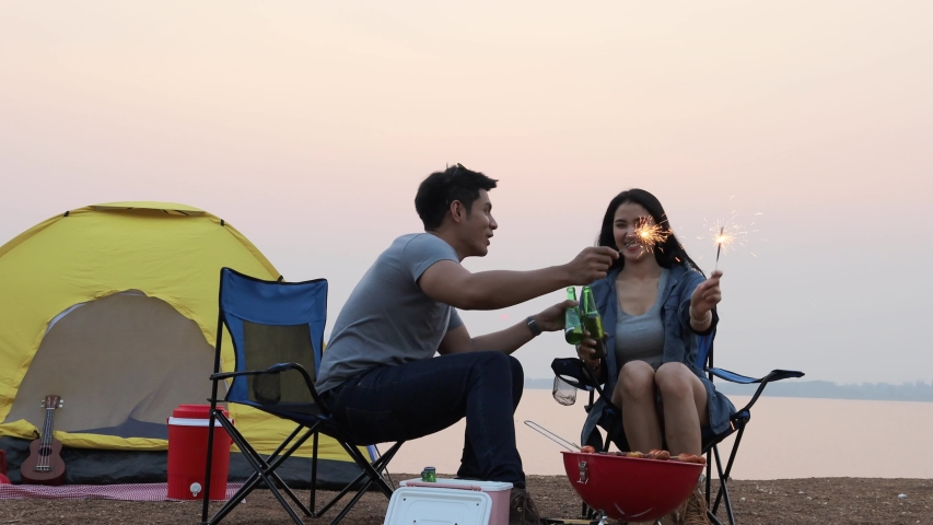 young asian couple drinking beer and playing sparkle together in front of tent at lakeside.