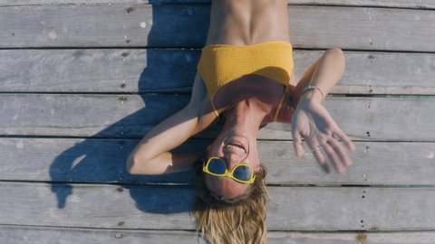 Aerial top down view, woman relaxing on wooden jetty over tropical water. Young attractive woman lying on her back on wooden dock. Aerial drone shot. Woman waving hello to camera 