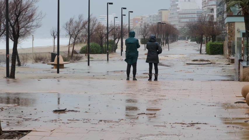 Storm Gloria in Spain. Big waves on Mediterranean Sea in Catalonia, Roses city. Consequence of storm, seafront is flooded with water, garbage. Pare of tourists in raincoats walking and makes photos. 