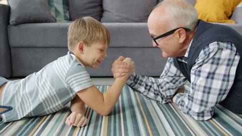 Elderly man and little boy are practising arm wresting at home enjoying activity, child is winning. Modern pastime, family relationship and fun concept.