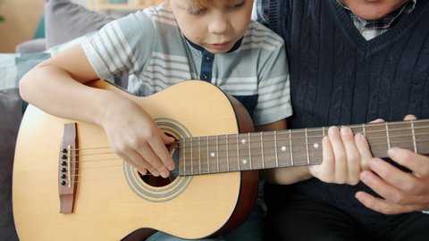 Caring grandfather is helping cute kid teaching guitar playing at home together enjoying domestic music lesson. People and musical education concept.