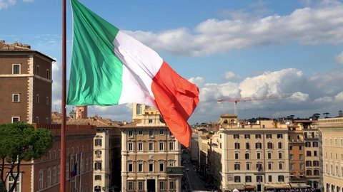 Flag of Italy fluttering in the wind over a square in Rome against a blue sky with clouds.