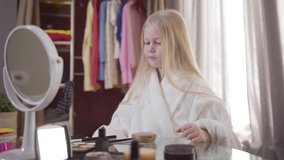Portrait of smiling blond Caucasian girl doing makeup indoors. Cute cheerful child applying face powder and eye shadows. Beauty, fashion, childhood.