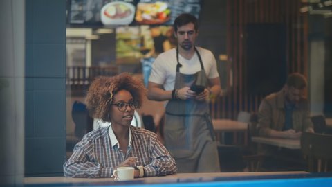 African-american young female customer keeping her wrist over electronic payment machine while paying for order through smartwatch in cafe. Waiter holding terminal while client paying with smartwatch