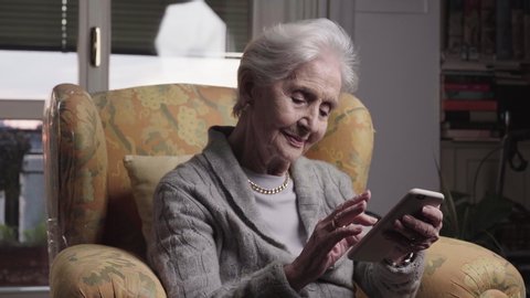 94 years old skilled lady portrait using smartphone technologies to communicate easily from her living room, senior woman sitting in yellow armchair holding phones and texting to family. 