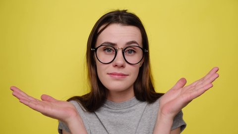 Dissatisfied woman imagining alternatives, weighs pros and cons, makes hard solution, spreads palms, cant choose answer, frowns face, wears spectacles and grey t-shirt, isolated over yellow background