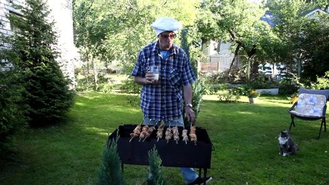 Senior  man is frying shish kebab in garden, drinking beer.  Cheerful funny manin chef  is frying food.  Sunny summer day. 
