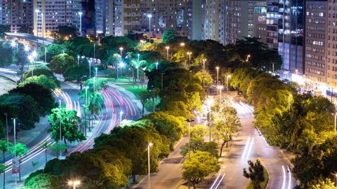 Aerial night timelapse of dense traffic on large street avenue in Rio de Janeiro, Brazil. View of cars passing on the road between trees, bright lights. Buildings in the background.