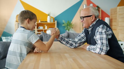 Happy child is enjoying arm wrestling with loving grandfather in apartment, people are exercising and boy is winning. Family sports and lifestyle concept.