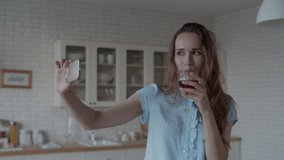 Close up young woman posing for photo with glass of red wine in kitchen interior in slow motion. Happy woman drinking wine in front of phone camera in kitchen. Pretty girl making video on smartphone.