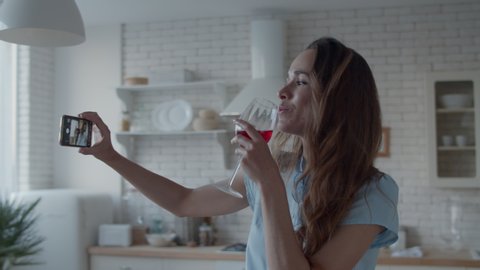 Close up young woman posing for photo with glass of red wine in kitchen interior in slow motion. Happy woman drinking wine in front of phone camera in kitchen. Pretty girl making video on smartphone.
