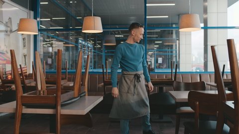 Young waiter or cafeteria owner putting chairs on tables to clean area at the end of workday. Male restaurant owner preparing in for opening. Small business and startup concept