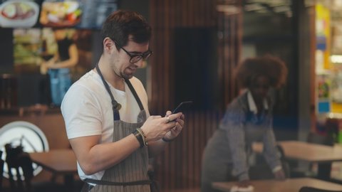 Portrait of caucasian barista guy or waiter wearing apron smiling and typing on mobile phone in cafe or coffeehouse. African waitress cleaning table on background. Waiter chatting online on phone