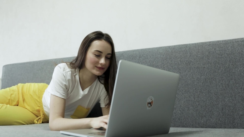 Smiling young woman sitting on couch using laptop notebook looking at screen typing message,