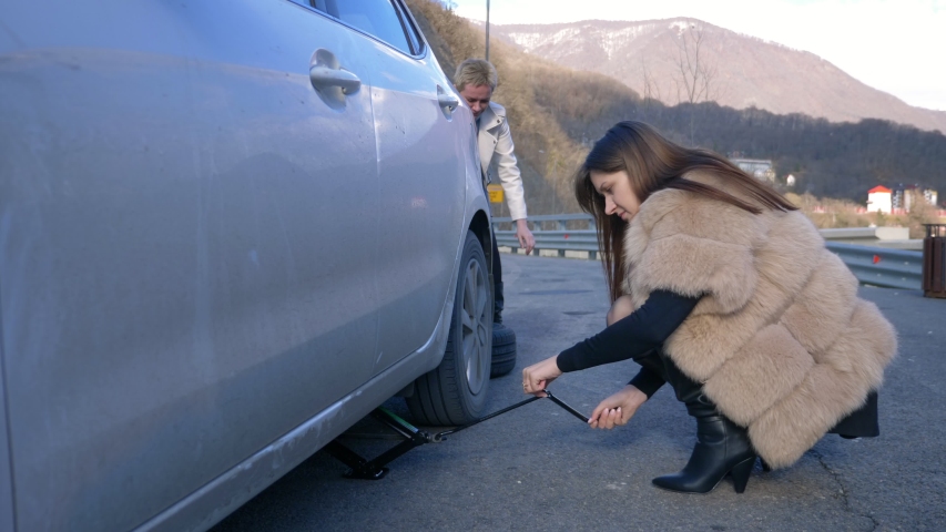two girls change a flat tire on the car on the side of the road
