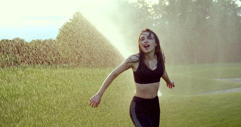 Emotional sunny portrait of the attractive young brunette having fun while dancing and spinning round in the rain from the sprinkler.