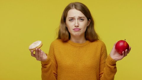 Beautiful slim girl in sweater holding apple and donut. making choice between healthy fruit and junk food, useful via harmful nutrition, diet concept. indoor studio shoot isolated on yellow background