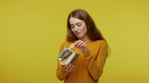Cute girl in casual sweater eating crisp chips with expression of pleasure. enjoying tasty fried food, unhealthy high calorie nutrition, diet concept. indoor studio shot isolated on yellow background