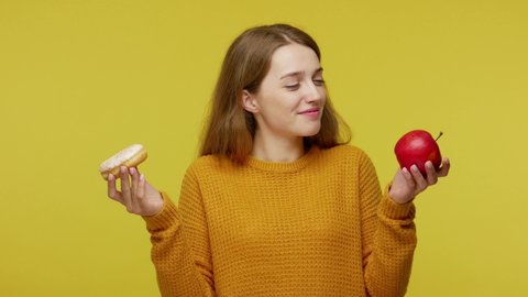 Attractive slim girl in sweater holding apple and donut. choosing and biting healthy fruit instead junk food, useful via harmful nutrition, diet. indoor studio shoot isolated on yellow background