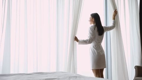Smiling attractive brunette woman wearing nightgown and robe, opening curtains lace, looking out of window, greeting new day. Happy beautiful young lady enjoying good morning free time at home.