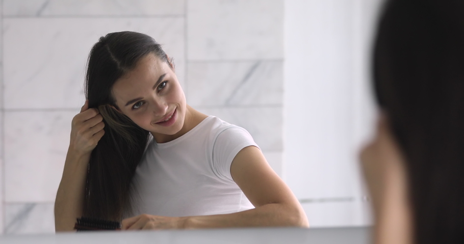 Attractive millennial lady looking in mirror, combing long healthy straight hair. Confident young brunette woman doing hairstyle in bathroom, getting ready in morning, enjoying everyday routine. | Shutterstock HD Video #1045279366
