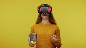 Young woman eating crisp chips and watching 3d mp4ie with VR glasses, smiling enjoying film, saying wow experiencing emotions in virtual reality. indoor studio shot isolated on yellow background