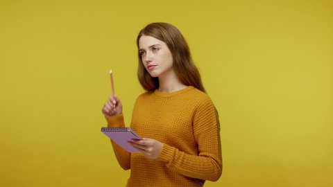 Cute girl in sweater standing with pensive expression and scratching head with pencil. writing down idea in notebook, making plans in organizer. indoor studio shot isolated on yellow background