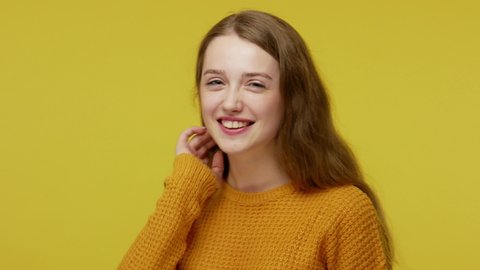 Beautiful good-natured girl in pullover with kind face expression and perfect smile looking playfully. winking at camera and adjusting her hair, flirting. studio shot isolated on yellow background
