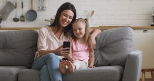 Joyful young mother embracing little preschool daughter, watching funny cartoons on smartphone, singing sound track songs, enjoying weekend time. Happy family using foreign language learning apps.