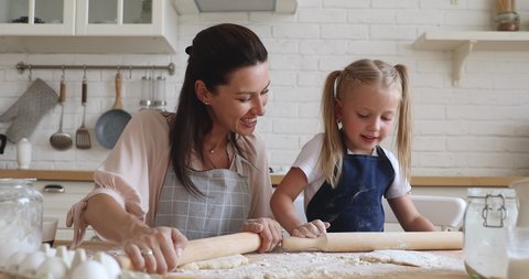 Happy 30s woman teaching little preschool daughter flattening dough. Smiling family of two preparing homemade bakery food together in modern kitchen. Pleasant mommy showing how to use rolling pin.
