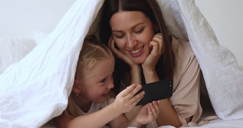 Happy 30s mother lying on bed under duvet with joyful little daughter, small kid girl holding cell phone, taking selfie with smiling mommy, using funny mobile apps, having fun together in bedroom.