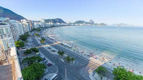 Day to night sunset timelapse of Copacabana skyline in Rio de Janeiro city in Brazil. View of the front street traffic with car road traffic. Busy ocean beach full of activity, chairs, umbrellas, and 