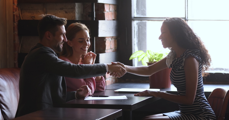 Happy young couple signing contract with african ethnicity realtor saleswoman wedding planner after discussing details in cafe. Satisfied smiling clients making agreement with mixed race professional. | Shutterstock HD Video #1045314478