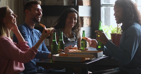 Happy african american guy holding bottle of beer and pizza slice, telling funny joke storytelling to laughing multiracial friends. Excited mixed race people enjoying conversation while sharing lunch.