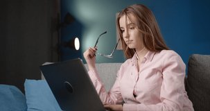 Thoughtful young lady with long hair putting on protective glasses for working with laptop while sitting on comfortable sofa. Charming female student preparing for exams at home with modern design