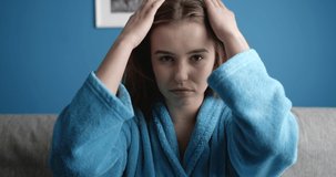 Portrait of charming brunette woman in azure colored bathrobe fixing her long straight hair after taking morning shower. Attractive girl sitting on grey couch with classic blue wall behind