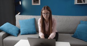 Concentrated female student in eyeglasses, rose blouse and black trousers using laptop for searching information. Young lady with long hair doing homework at home with trendy colored interior