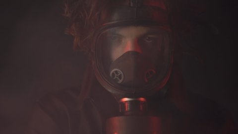 Young man in gas mask and black clothes among poison smoke on dark background