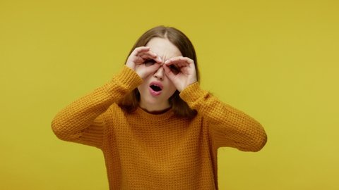 Cute inquisitive girl in casual clothes looking around through fingers imitating binoculars, saying wow, being surprised amazed by what discovered. indoor studio shot isolated on yellow background