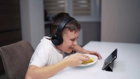 Boy eating at home while looking at digital tablet. Child in headphones sitting at the table in the kitchen and eating. Gadgets and children.