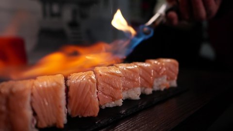 a process of preparation of sushi is Philadelphia, burning of smoking fire of fresh raw red fish salmon tuna eel, on a black background before a serve to the clients