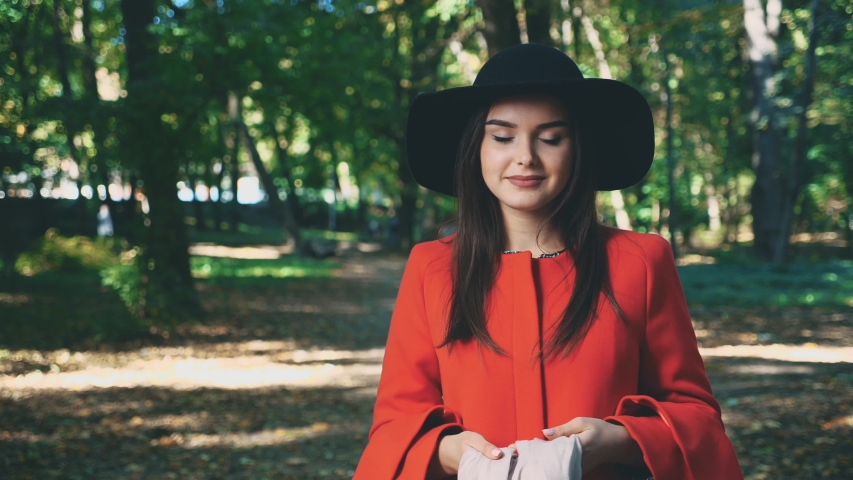 Pretty young girl with red coat and black hat is binding gently-pink scarf, isolated, on park blurred background. Close up. Copy space. 4K.